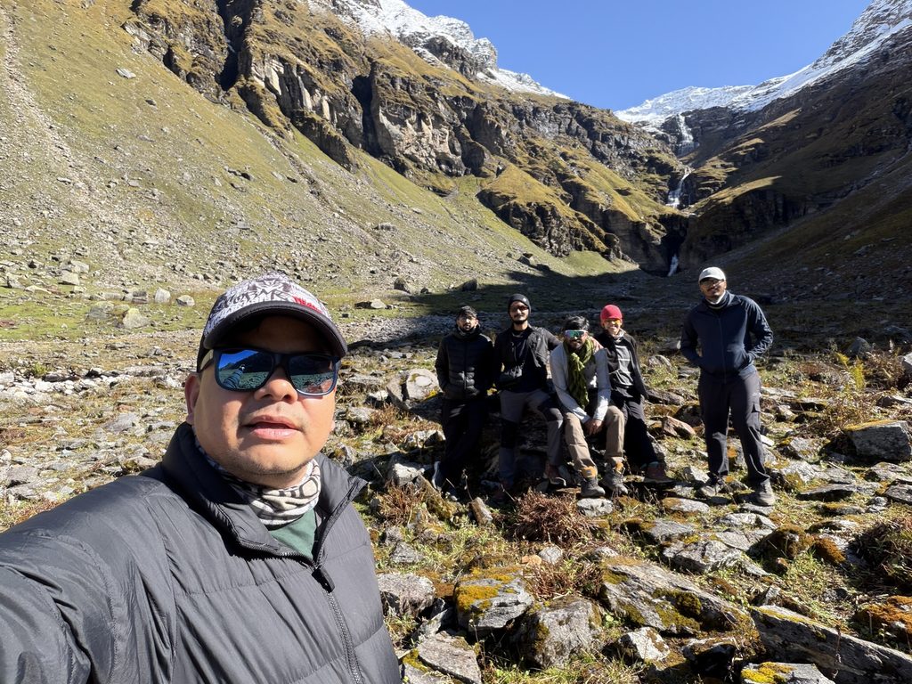 Trekker group posing in a rocky Himalayan valley beneath snow-covered peaks on the Rupin Pass trek